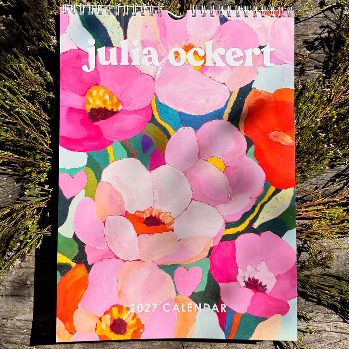 A spiral-bound 2027 Julia Ockert calendar featuring a vibrant floral cover design, placed on a rustic weathered wooden surface surrounded by green foliage.
